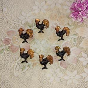 May include: Six gold and black rooster figurines on a beige background with a floral pattern. The roosters are facing different directions.