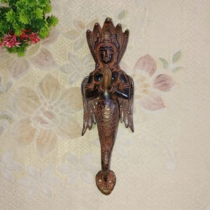 May include: A brass door handle shaped like a woman with wings and a snake head. The handle is dark brown with gold accents.