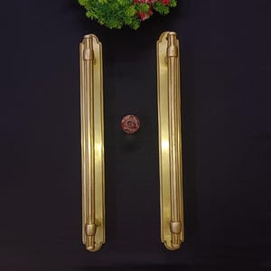 May include: Two long, brass-colored door handles with vertical grooves and decorative ends. A small, round, red and brown knob is centered between the handles. The handles are set against a dark background.