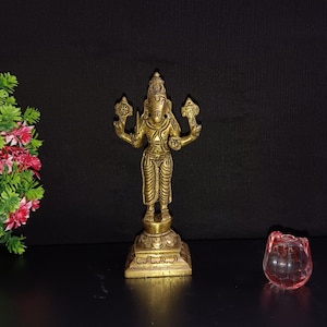 May include: A gold-colored brass statue of a Hindu deity with four arms, standing on a square base. The deity is holding various objects in its hands, including a conch shell and a lotus flower.