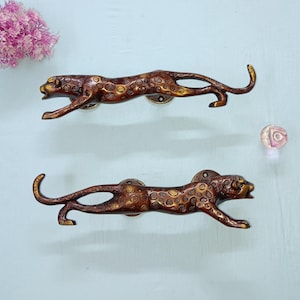 Peut inclure: Deux poignées de meuble en forme de léopard de couleur bronze. Les poignées présentent des détails de taches et sont en position de course. Le matériel est monté sur une surface bleu clair, avec un arrangement floral rose et une perle de verre décorative.