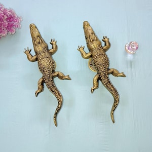 May include: Two metallic gold-toned door handles shaped like crocodiles. Each handle features a textured, scaly surface, a long tail, and four legs extended outward. The crocodile figures are positioned vertically, mirroring each other in design.
