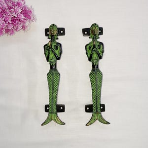 May include: Two decorative mermaid-shaped door handles. The handles are green and black with a detailed design. The mermaid figures have their hands clasped in front of them. The handles are mounted on black rectangular plates.