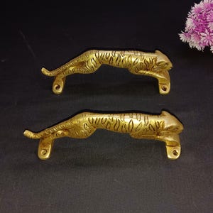 Può includere: Due maniglie per porte dorate a forma di tigri che saltano. Le maniglie presentano un motivo a tigre dettagliato e sono montate su una superficie nera.