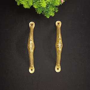 May include: Two gold-toned door handles with a decorative design featuring a cross and a face. The handles are made of metal and have a polished finish.