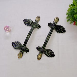 Op de afbeelding: Twee decoratieve zwarte en gouden deurgrepen met een bladachtig ontwerp. De handgrepen hebben een donkere afwerking met groene accenten en sierlijke goudkleurige details. De handgrepen zijn waarschijnlijk voor een kast of deur.