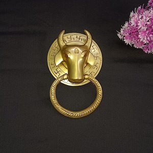Puede incluir: Un llamador de puerta de color dorado con forma de cabeza de toro, con un diseño circular que presenta un patrón repetido del símbolo hindú 'Swastika'. El llamador está unido a un anillo.