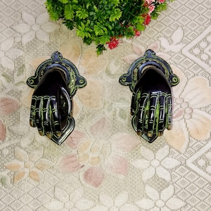 May include: Two black and green hand-shaped door knockers with intricate details. The knockers are mounted on a white background with a floral pattern.