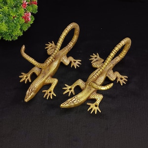 Pode incluir: Dois ganchos de parede em forma de lagarto de metal dourado. Os lagartos estão virados em direções opostas e têm escamas e garras detalhadas.