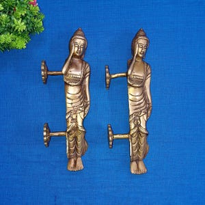 Peut inclure: Une paire de poignées de porte dorées avec un design détaillé de deux figures de Bouddha en position debout. Les poignées sont fixées à une base métallique avec un motif de rouleau décoratif.