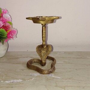 May include: A gold brass incense burner shaped like a cobra with a round top. The burner is on a white surface.