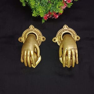 Pode incluir: Um par de batentes de porta ornamentados em tom dourado, em forma de mãos, cada um com um design detalhado e um anel para bater.