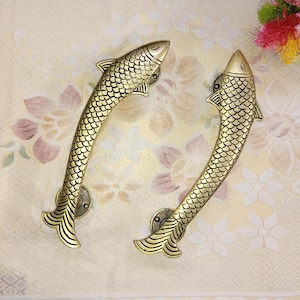 May include: Two gold-toned fish-shaped door handles with a detailed, scaly design. The handles are curved and have a decorative fin at the end.