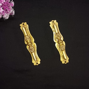 May include: Two gold-toned metal door handles with ornate designs. The handles are rectangular with a decorative, raised, central section.