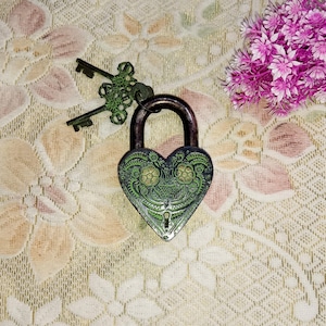 May include: A green heart-shaped padlock with intricate floral designs and a keyhole. The padlock is on a white and pink floral patterned surface.