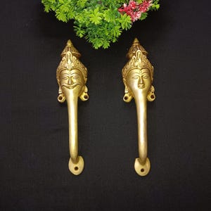 Könnte beinhalten: Zwei goldfarbene Türklinken mit verzierten Designs, die ein Gesicht mit einer Krone zeigen. Die Griffe befinden sich auf einem schwarzen Hintergrund.