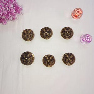 May include: Six gold-toned metal buttons with a raised design of a face. The buttons are arranged in two rows of three on a white surface.