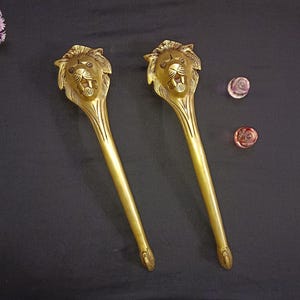 Puede incluir: Dos manijas decorativas doradas con cabeza de león. Las manijas tienen un diseño largo y delgado con una cabeza de león en la parte superior. La cabeza de león tiene detalles. También se ven dos pequeños objetos decorativos redondos rosas y rojos.