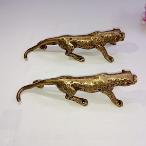 Op de afbeelding: Twee gouden metalen leeuwinfiguren. De leeuwinnen zitten in een hurkende positie met hun hoofd naar de zijkant gedraaid. De figuren zijn gedetailleerd met vlekken en hebben een realistisch uiterlijk.
