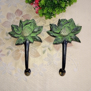 Puede incluir: Dos ganchos de metal negro con diseños de flores de loto verdes. Los ganchos son decorativos y se pueden usar para colgar toallas, abrigos u otros artículos.