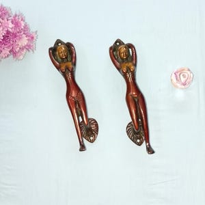 May include: Two bronze-colored door handles in the shape of a woman with arms raised above her head. The handles have a reddish-brown finish and are mounted on a light blue surface. A cluster of pink flowers is visible.