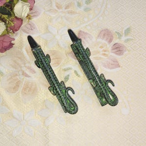 Peut inclure: Deux butées de porte en métal vert et or en forme de lézard avec un design détaillé. Les butées de porte en forme de lézard sont sur un fond blanc à motif floral.