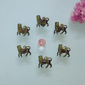 Puede incluir: Seis figuras de leones de color bronce con detalles intrincados están dispuestas en círculo alrededor de una pequeña esfera de vidrio iridiscente. Los leones tienen un acabado verde y marrón. Un racimo de flores rosas está en la esquina superior derecha.