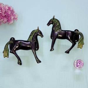Może przedstawiać: Dwa ciemnobrązowe haczyki ścienne w kształcie konia z zielonymi i złotymi akcentami. Konie stoją, ze szczegółowymi grzywami i ogonami. Różowy kwiat i dekoracyjna szklana kula również znajdują się na zdjęciu.