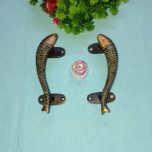 Puede incluir: Un par de tiradores de puerta de latón antiguo con forma de pez con detalles en negro y dorado. Los tiradores están montados sobre un fondo azul claro.