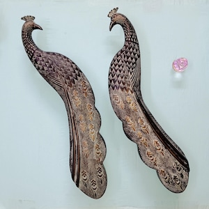 Puede incluir: Dos colgantes de pared decorativos de pavo real. Las aves son marrones con patrones de plumas detallados en oro y marrón. Los pavos reales tienen una corona en la cabeza y están colocados sobre un fondo azul claro. También se ven una flor rosa y una pequeña gema rosa.