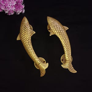 May include: A pair of gold-toned fish-shaped door handles. The handles are detailed with scales and have a curved shape.