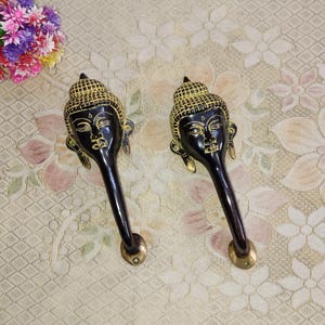 Op de afbeelding: Twee zwarte en gouden metalen deurgrepen met een Boeddha-gezichtsontwerp. De grepen zijn lang en gebogen, met een decoratieve knop aan de onderkant.