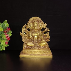 Puede incluir: Una estatua de metal dorado de una deidad hindú con cuatro cabezas y cuatro brazos. La deidad está sentada en un trono con una base decorativa.