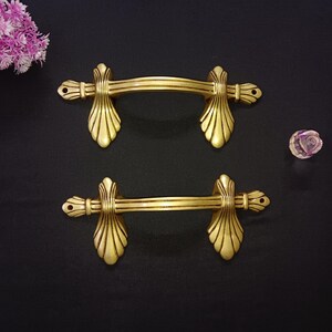 Op de afbeelding: Twee antieke gouden kastgrepen. Elke greep heeft een gebogen stang en decoratieve uiteinden. De grepen staan tegen een donkere achtergrond, met een kleine decoratieve bloem in de hoek. Ze zijn waarschijnlijk van metaal.