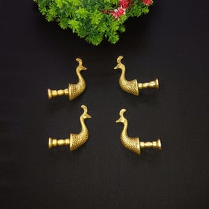 May include: Four gold-colored peacock-shaped drawer pulls arranged on a black surface. Each pull features a detailed peacock design with a long neck and a decorative tail. Green and red floral accents are in the top corner.