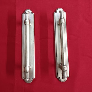 Puede incluir: Dos tiradores de puerta plateados con una placa rectangular y una barra vertical. Los tiradores tienen un diseño decorativo con la parte superior e inferior redondeadas.