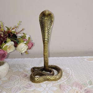 May include: A detailed brass cobra statue with a raised hood and coiled body. The snake is a golden color, with intricate scale detailing. The statue is displayed on a patterned tablecloth, with a vase of flowers in the background.