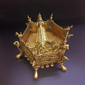 Può includere: Una lampada a olio esagonale in ottone con dettagli intricati e una figura appesa. La lampada ha quattro gambe a forma di uccello e una catena decorativa.