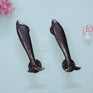 May include: Two bronze-coloured fish-shaped door handles. The handles have a detailed scale pattern and are mounted on round plates. The handles are decorative and add a unique touch to any door or cabinet.