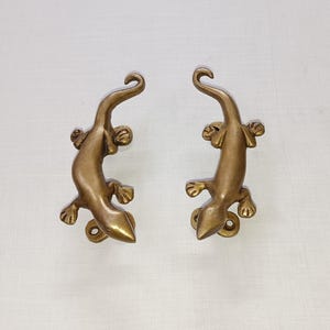 以下が含まれることがあります： ブロンズ色のトカゲ型の装飾ドアプル2つ。各トカゲは、丸まった尾、4つの詳細な足、テクスチャード加工された体を持っています。ドアプルは白い表面に取り付けられています。