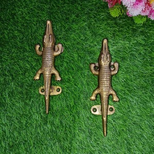 May include: Two gold-toned crocodile-shaped door handles. The handles are detailed with textured scales and have a realistic appearance.