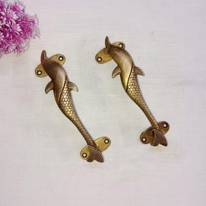 Könnte beinhalten: Zwei goldfarbene, delphinförmige Türklinken mit detaillierten Schuppen. Die Griffe sind aus Metall und haben eine polierte Oberfläche.