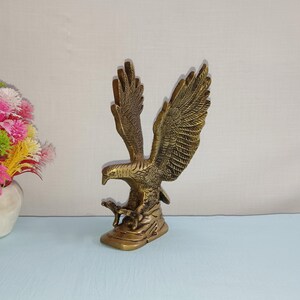 May include: A gold-colored metal eagle sculpture with its wings spread wide. The eagle is perched on a base with a smaller eagle sculpture underneath it.