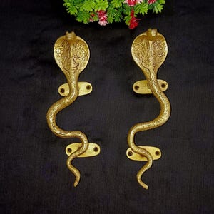 May include: A pair of gold-toned door handles shaped like cobras with intricate details. The handles are mounted on a rectangular base with two screws.