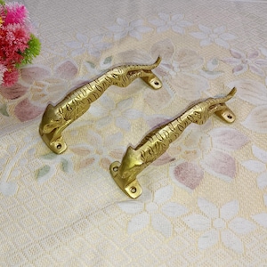 Può includere: Due maniglie per porte color ottone, ciascuna a forma di tigre accovacciata. Le maniglie presentano intagli dettagliati e sono montate su basi rettangolari con fori per le viti. Lo sfondo è un tessuto color crema con un motivo floreale.