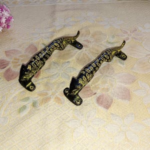 Può includere: Due maniglie per porte in metallo nero a forma di pantere stilizzate con accenti dorati. Le maniglie sono su uno sfondo bianco.