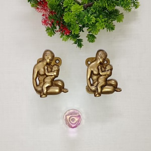 Puede incluir: Dos esculturas de metal dorado de una madre y un niño, con un diseño de ojo de cerradura para colgar. Un pequeño objeto de vidrio redondo, rosa y morado está debajo. Follaje artificial verde y rojo en la parte superior.