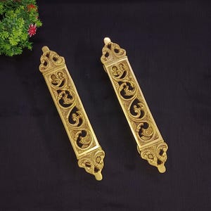 Może przedstawiać: Dwa ozdobne złote metalowe uchwyty do drzwi z misternymi wzorami kwiatowymi. Uchwyty mają kształt prostokątny i ozdobne górne i dolne części.