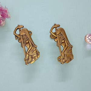 May include: Two antique brass door pulls, each depicting a woman in a flowing gown. The pulls are a warm gold colour and are mounted on a light blue surface. A pink flower arrangement is in the upper left corner.