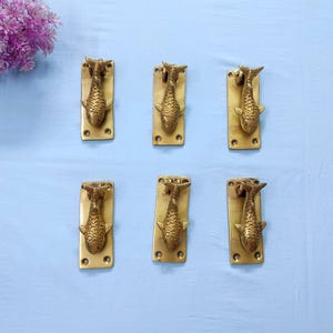 May include: A set of six brass door knockers, each featuring a fish-shaped design mounted on a rectangular brass plate with four screw holes. The metallic finish is polished and gold-toned, showcasing detailed scales and fins on each fish figure.